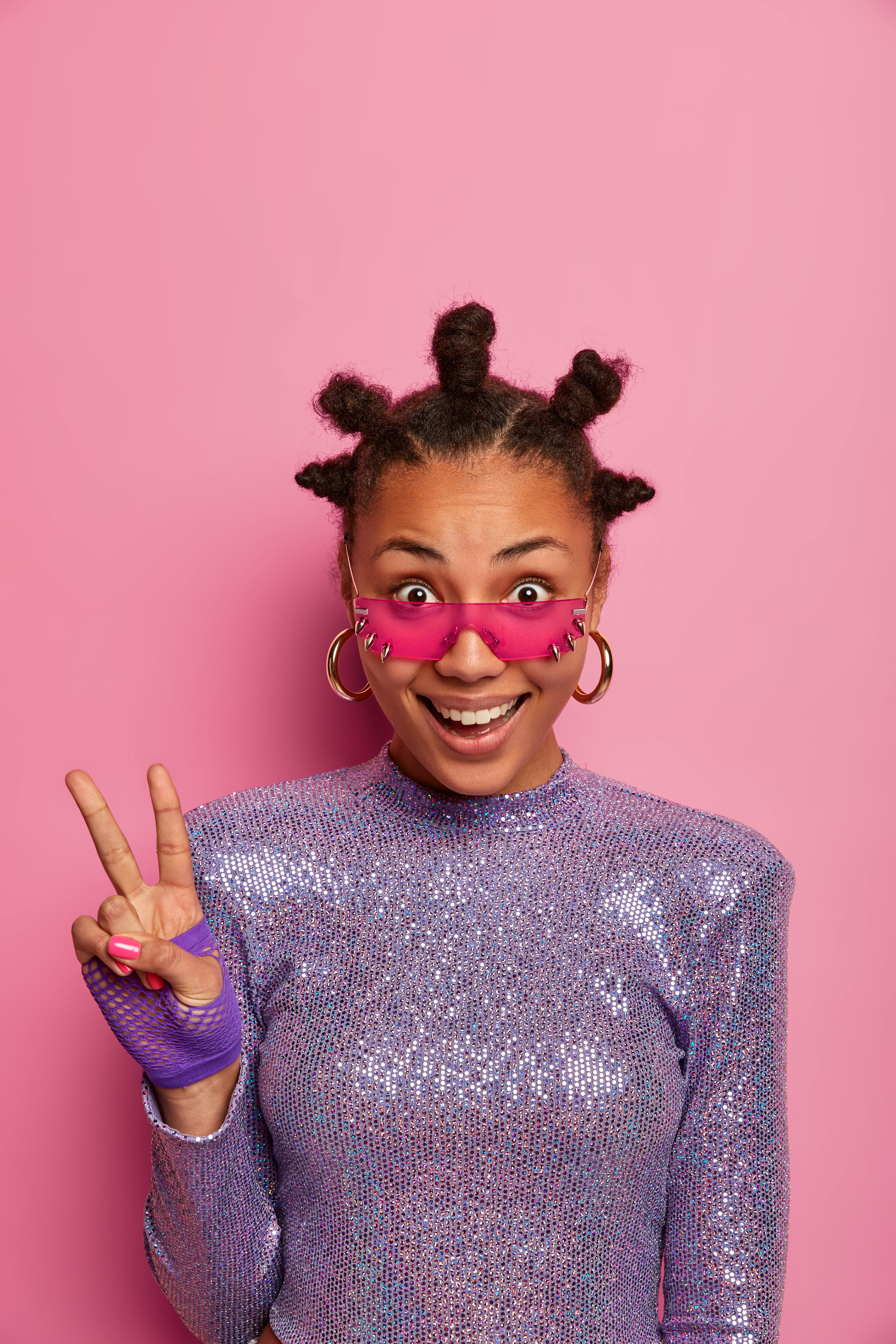 Pretty joyful dark skinned woman with bun hairstyle shows peace gesture looks through pink sunglasses follows fashion trends stays always positive models over pink wall copy space above