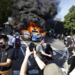 Protesters surround burning car in street demonstration.
