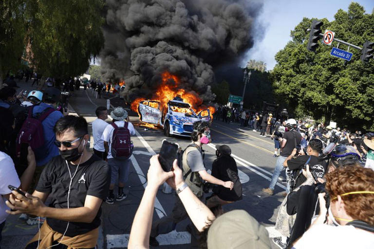 Protesters surround burning car in street demonstration.