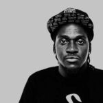 Man in cap and black shirt, grey background.