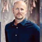 Man in blue shirt stands against blurred background.