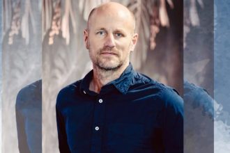 Man in blue shirt stands against blurred background.