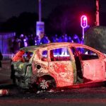 Burnt car on street at night, people observing.