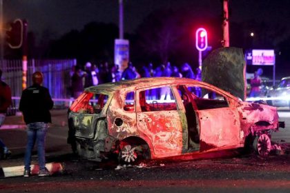 Burnt car on street at night, people observing.
