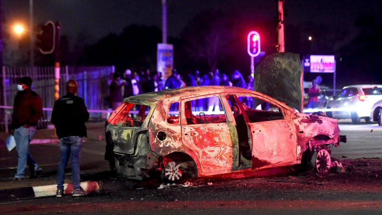 Burnt car on street at night, people observing.