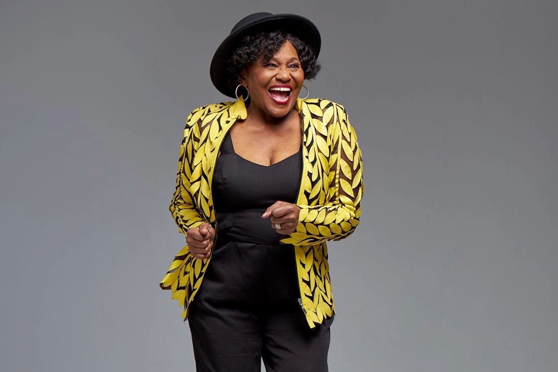 Smiling woman in yellow patterned jacket, black hat.