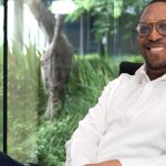 Smiling person sitting in office with greenery outside.