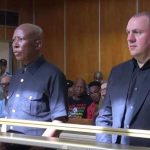 Two men standing in courtroom with audience behind.