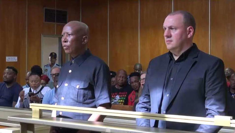 Two men standing in courtroom with audience behind.