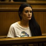 Woman seated in courtroom, wearing white shirt.