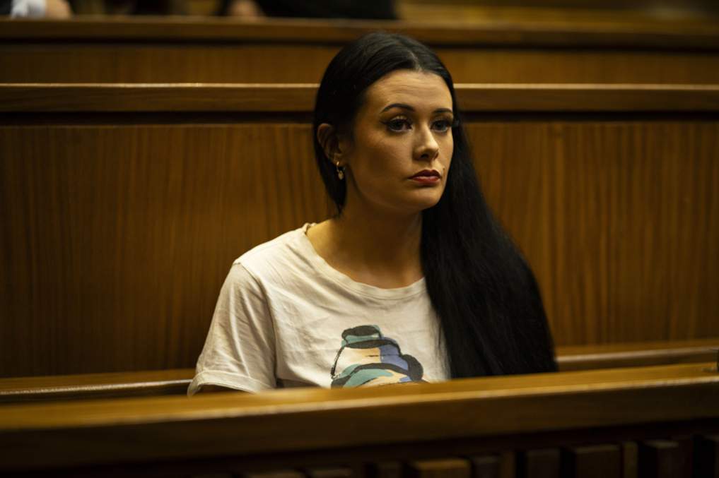 Woman seated in courtroom, wearing white shirt.