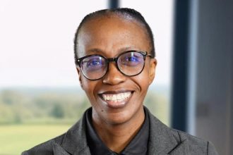 Person smiling, wearing glasses, grey suit indoors.