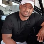 Man sitting in car, smiling and wearing cap.