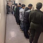 People lined up against wall, guarded by officer.