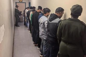 People lined up against wall, guarded by officer.