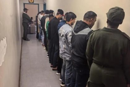 People lined up against wall, guarded by officer.