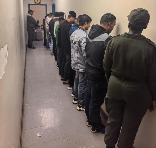 People lined up against wall, guarded by officer.