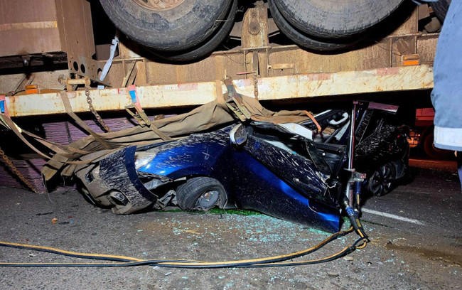 Blue car crushed under large truck trailer