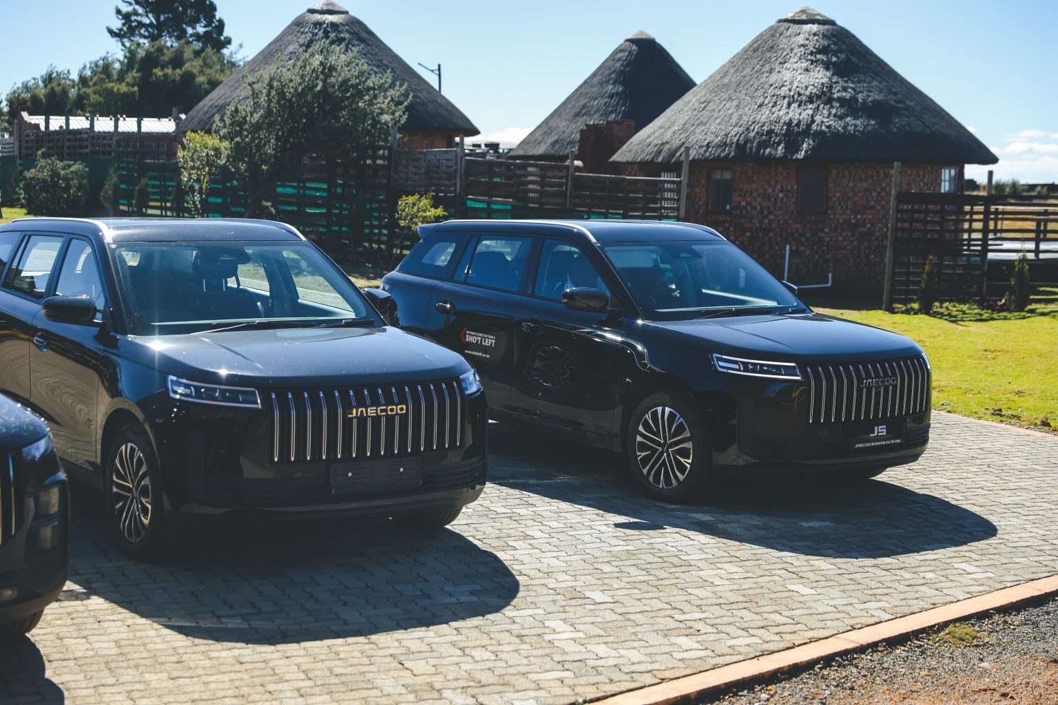 Two black Jaecoo SUVs parked near thatched lodges