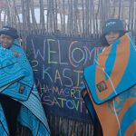 Two women wrapped in blankets beside welcome sign