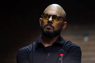 Bald bearded man wearing yellow-tinted glasses at microphone
